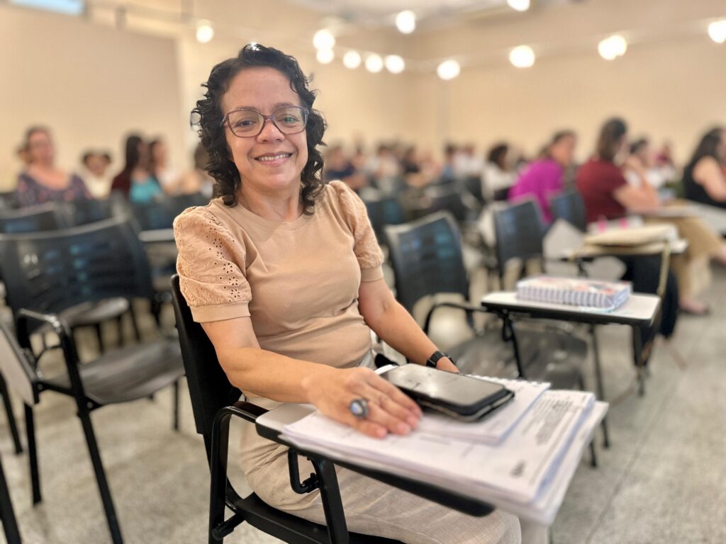 A mulher está sentada em uma sala de aula, com outras pessoas ao fundo. Ela está sorrindo para a câmera e usando óculos. Sua roupa é uma blusa bege com mangas curtas e bufantes, e uma calça bege clara. Em sua mão direita, ela tem um celular em cima de alguns papéis. Há um anel azul em seu dedo. A sala de aula tem carteiras com mesas acopladas e iluminação no teto.