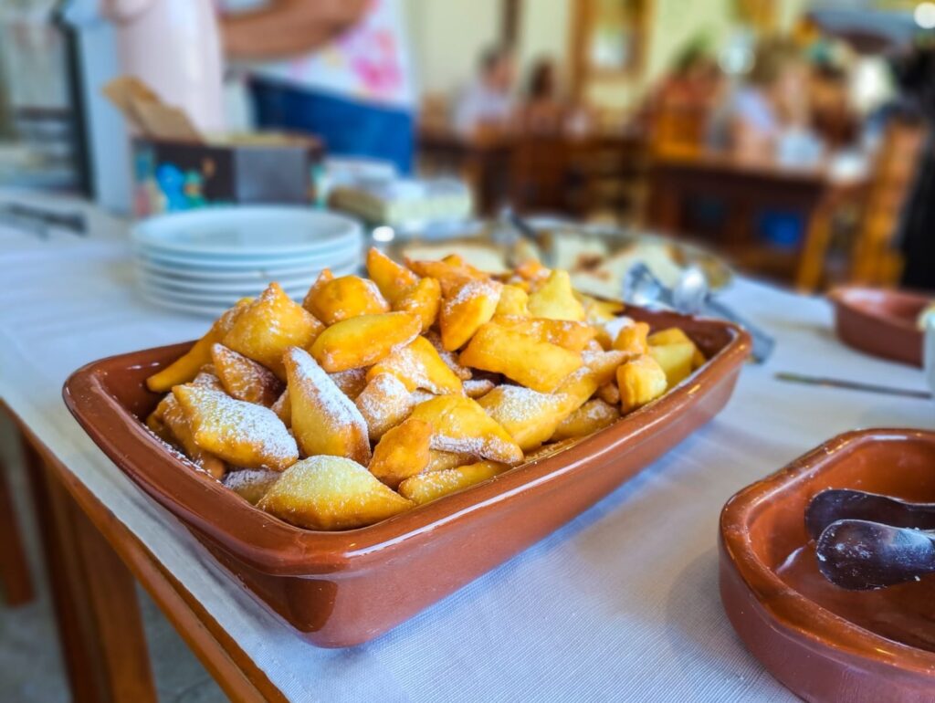 A imagem mostra uma travessa retangular de barro, cheia de pequenos doces fritos polvilhados com açúcar de confeiteiro. Esses doces têm uma cor dourada e parecem ser feitos de massa frita, com formatos variados, alguns mais arredondados e outros mais pontiagudos. Eles estão dispostos de forma a preencher a travessa, com alguns pedaços empilhados sobre outros. Ao lado da travessa principal, há outra travessa menor de barro, onde se vê um par de pinças. O fundo da imagem está desfocado, mas é possível notar uma toalha branca sobre uma mesa, e ao fundo, pessoas sentadas em mesas, sugerindo um ambiente de restaurante ou buffet. Há também uma pilha de pratos brancos em segundo plano, à esquerda. A iluminação parece ser natural, vinda de cima ou da frente, realçando o brilho dos doces.