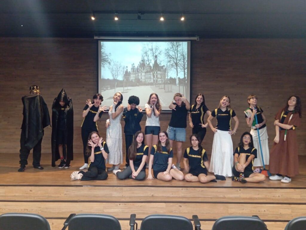 A imagem mostra um grupo de jovens em um palco, possivelmente durante uma apresentação teatral ou evento escolar. Eles estão dispostos em duas fileiras, com alguns em pé e outros sentados no chão. Ao fundo, há uma tela projetando uma imagem de um castelo e árvores. Na fileira de trás, da esquerda para a direita, vemos dois jovens vestidos com trajes escuros e capuzes, um deles com uma coroa dourada. Em seguida, há um grupo de jovens com roupas mais casuais, alguns fazendo poses divertidas para a câmera. Uma jovem está vestida com um longo vestido branco, e outra usa um vestido marrom. Na fileira da frente, há jovens sentados, alguns usando camisetas pretas com a inscrição "OBJETIVO" em amarelo. As expressões e poses sugerem um ambiente descontraído e animado. A iluminação do palco e o fundo de madeira criam um cenário adequado para uma performance.