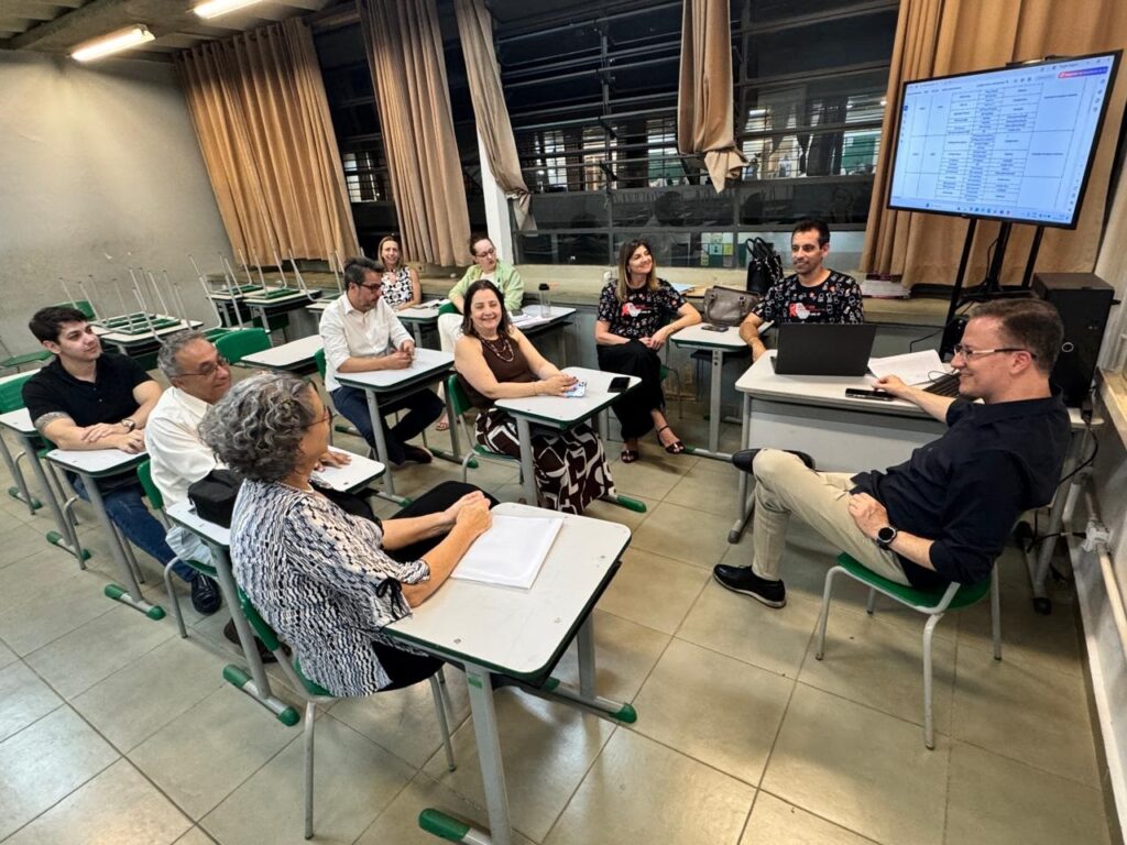 A imagem mostra uma sala de aula com várias pessoas sentadas em carteiras organizadas em filas. A maioria está voltada para a frente da sala, onde há uma grande tela exibindo algum tipo de tabela ou planilha. As pessoas parecem ser alunos e um professor ou instrutor, que está à direita, sentado e sorrindo, vestindo camisa azul escura e calça clara. A sala é iluminada por luz artificial e tem paredes claras com janelas cobertas por cortinas. Há cadeiras empilhadas ao fundo e o chão é de azulejo. A atmosfera parece ser informal e descontraída, possivelmente durante uma aula, reunião ou apresentação.
