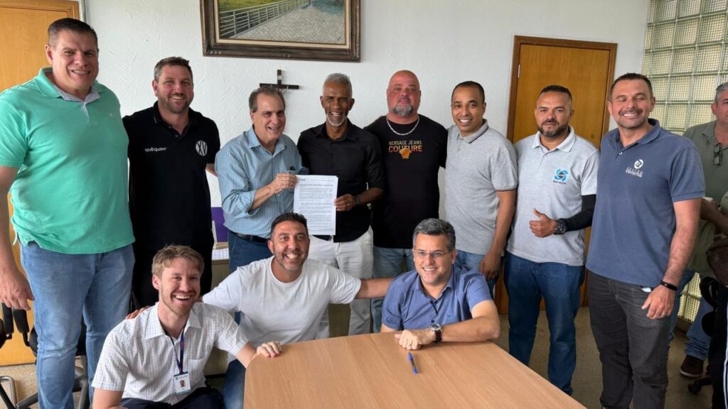 A imagem mostra um grupo de 11 homens em um ambiente interno, possivelmente um escritório. Eles estão posando para uma foto, alguns em pé e outros sentados. Na parte superior esquerda, um homem de camisa polo verde e jeans sorri para a câmera. Ao lado dele, um homem de camisa preta com um logo e jeans também sorri. No fundo, um grupo de homens está em pé. Um deles, de camisa xadrez azul clara, segura um documento. Ao lado dele, um homem de camisa preta e um homem de camisa preta com a estampa "VERSACE JEANS COUTURE". Mais à direita, um homem de camisa cinza e outro de camisa azul escura completam a fileira. Na parte inferior da imagem, dois homens estão sentados à frente de uma mesa de madeira. Um deles, de camisa branca, sorri amplamente, enquanto o outro, de camisa azul, também sorri. Um terceiro homem, de camisa xadrez branca e azul, está sentado entre eles, sorrindo e com a mão apoiada no ombro do homem de camisa branca.