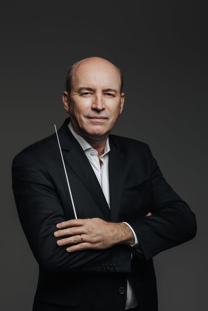 Um homem de meia-idade, careca, veste um terno preto com uma camisa branca aberta. Ele tem os braços cruzados e segura uma batuta na mão esquerda. Seus olhos olham diretamente para a câmera com uma expressão séria. O fundo é um cinza escuro e sólido. Há uma aliança dourada em seu dedo anelar esquerdo.