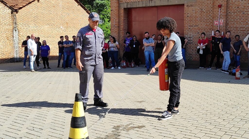A imagem mostra uma demonstração de uso de extintor de incêndio. Um bombeiro, vestindo uniforme cinza e boné, observa atentamente enquanto uma mulher com cabelo cacheado, vestindo camiseta azul e calça preta, utiliza um extintor vermelho. Ela direciona o jato do extintor para um cone de sinalização amarelo e preto, posicionado no chão de paralelepípedos. Ao fundo, um grupo de pessoas observa a demonstração. Algumas delas estão em pé, vestindo roupas casuais, enquanto outras parecem estar em uma área de treinamento, com vários extintores de incêndio vermelhos alinhados. A cena ocorre em um ambiente externo, com uma parede de tijolos vermelhos ao fundo e uma edificação com telhado aparente. A iluminação sugere que é um dia ensolarado.
