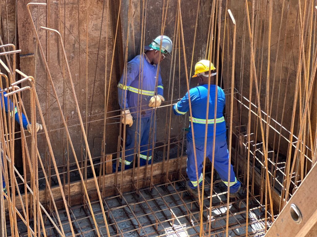 A imagem mostra dois trabalhadores da construção civil em um canteiro de obras, cercados por uma estrutura de vergalhões de aço. Ambos vestem uniformes de trabalho azuis e capacetes, um azul e outro amarelo, e luvas. Um dos trabalhadores, com capacete azul, está de frente para o outro, que usa capacete amarelo. Eles parecem estar inspecionando ou trabalhando nos vergalhões, que compõem uma malha complexa e vertical. O ambiente é rústico, com paredes de madeira e metal enferrujado ao redor, sugerindo que estão dentro de uma forma para concreto ou em uma área de fundação. A luz do sol ilumina a cena, criando sombras e realçando a textura dos materiais.