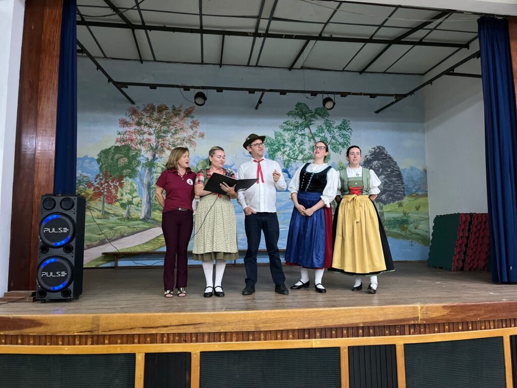 A imagem mostra cinco pessoas em um palco com um cenário pintado ao fundo. O cenário representa uma paisagem com árvores, montanhas e um rio. As pessoas estão vestidas de forma que sugere uma apresentação cultural ou folclórica. À esquerda, há uma caixa de som preta com luzes azuis acesas, com a marca "PULSE" visível. As cinco pessoas estão dispostas no palco, da esquerda para a direita: 1. Uma mulher com uma blusa vinho e calças vinho. 2. Uma mulher com um vestido estampado em tons de bege e verde, segurando um caderno aberto. Ela tem um lenço xadrez no pescoço. 3. Um homem com um chapéu marrom, camisa branca, gravata vermelha e calças escuras. Ele parece estar falando ou cantando. 4. Uma mulher com um vestido tradicional, com corpete preto sobre uma blusa branca de mangas bufantes, e uma saia azul longa. 5. Uma mulher com um vestido tradicional semelhante, com um corpete verde sobre uma blusa branca, e uma saia longa amarela. As roupas das mulheres 4 e 5 parecem ser trajes folclóricos típicos de alguma região da Europa, possivelmente da Baviera ou Áustria, com seus corpete e saias volumosas. O palco é de madeira, com uma estrutura aparente na parte inferior. Ao fundo, atrás das pessoas, o cenário pintado cria uma atmosfera rústica e campestre. Há cortinas azuis escuras nas laterais do palco.