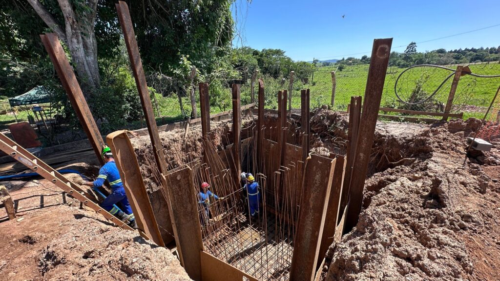 A imagem mostra uma vala profunda sendo escavada em um local rural, com trabalhadores da construção civil no fundo. A vala é reforçada com longas chapas de metal verticais, que parecem ser de aço, para evitar o desmoronamento das paredes de terra. No interior da vala, há uma estrutura de vergalhões de aço, indicando que uma fundação de concreto está em processo de construção. Dois trabalhadores, vestidos com uniformes azuis e capacetes, estão dentro da vala, um deles trabalhando na estrutura de vergalhões e o outro supervisionando. Na lateral da vala, outro trabalhador, também de uniforme azul e capacete verde, está subindo uma escada de madeira. Ao redor da vala, o terreno é de terra batida e há vegetação, com árvores e um campo verde ao fundo, cercado por uma cerca de arame. O céu está limpo e azul, sugerindo um dia ensolarado. A cena geral retrata um projeto de construção em andamento em um ambiente natural.