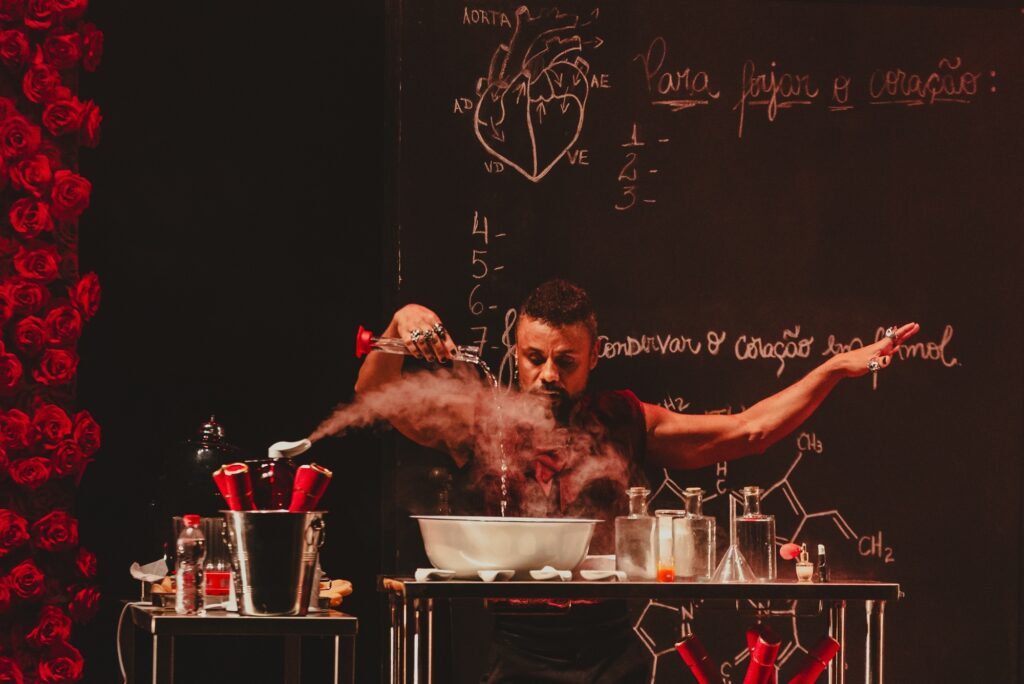A imagem retrata uma cena teatral dramática com um homem no centro, realizando um experimento. Ele está em um palco com um fundo preto, onde estão desenhados em giz um diagrama do coração humano e fórmulas químicas. À esquerda, uma cascata de rosas vermelhas artificiais desce pelo palco, adicionando um toque de cor e simbolismo à cena. O homem, vestindo uma roupa escura com detalhes vermelhos, está de frente para uma bancada onde se encontram diversos materiais de laboratório, como béqueres, frascos, um balde de metal com tubos vermelhos e uma tigela branca. Ele segura um tubo de ensaio de vidro com uma substância borbulhante, de onde sai uma fumaça densa que se espalha pelo ar. Sua expressão é intensa e focada, transmitindo a seriedade do momento. A iluminação foca no homem e em seus experimentos, criando um ambiente misterioso e envolvente. Ao fundo, a escrita em giz sugere um tema relacionado à preservação do coração, possivelmente em álcool, e instruções para "fazer o coração".