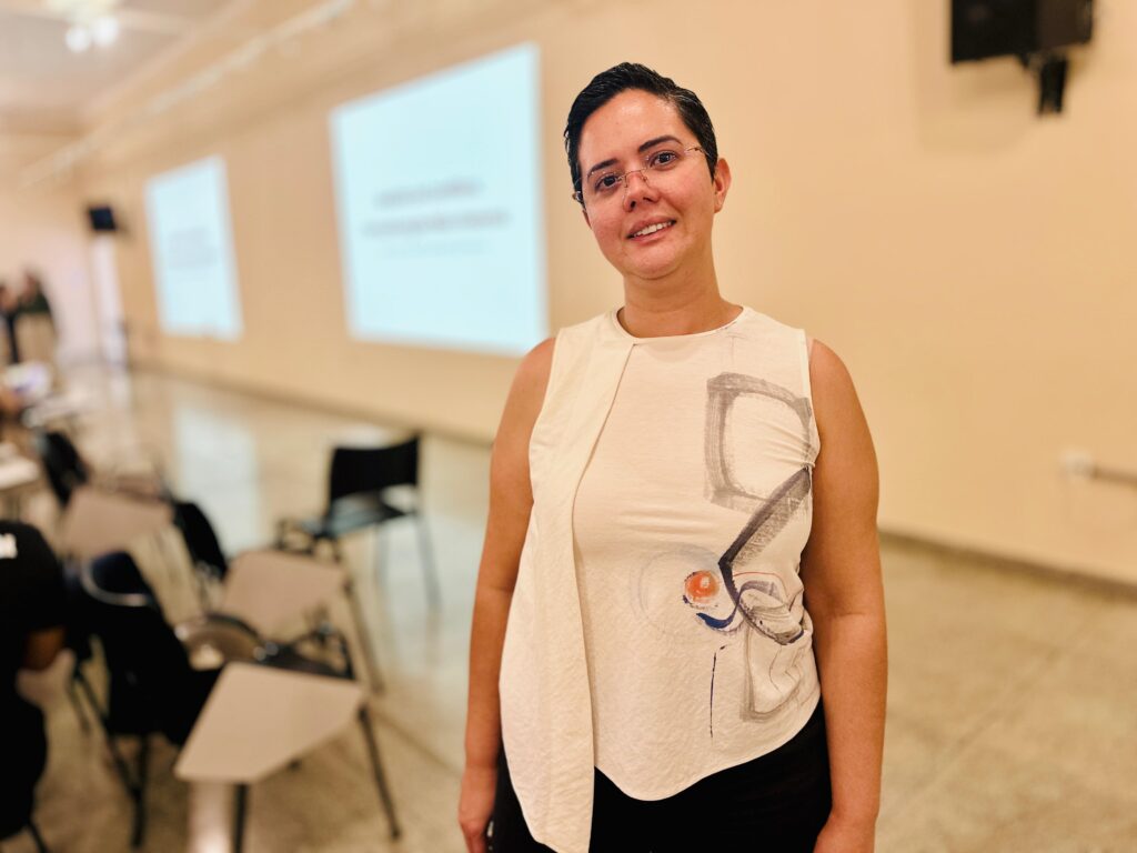 A mulher com cabelos escuros curtos e óculos está sorrindo para a câmera. Ela está usando uma blusa branca sem mangas com um design abstrato em cinza, laranja e azul. A blusa tem uma dobra assimétrica no lado esquerdo. Ela está em uma sala com cadeiras e mesas dispostas, e projeções em uma parede ao fundo.