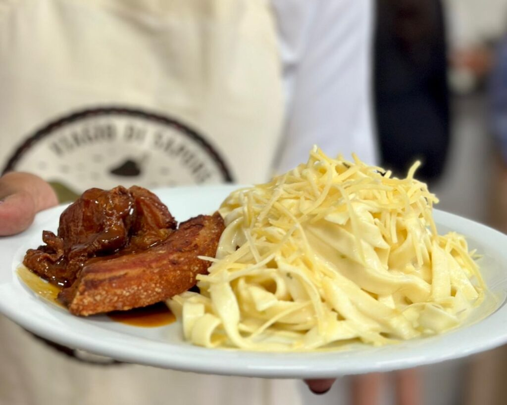 A imagem mostra um prato branco com uma porção de carne de porco com molho escuro e crocante ao lado de uma porção de massa com queijo ralado por cima. A carne parece ser barriga de porco, com a pele bem dourada e crocante, e a parte carnuda está coberta por um molho brilhante e espesso. A massa é longa e plana, possivelmente fettuccine, envolta em um molho cremoso e coberta com uma generosa quantidade de queijo ralado, que parece ser parmesão ou um queijo similar. O prato está sendo segurado por uma pessoa, e ao fundo, desfocado, é possível ver um avental com um logotipo circular. A iluminação destaca a textura apetitosa da comida.