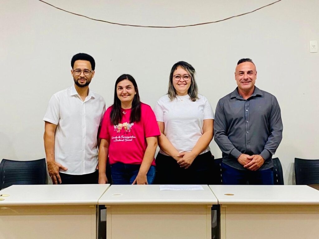 A imagem mostra quatro pessoas em pé atrás de uma mesa, com um fundo branco. Da esquerda para a direita, há um homem com óculos, camisa branca e calças escuras; uma mulher com uma camiseta rosa vibrante e jeans; uma mulher com uma camiseta branca e calças escuras; e um homem com uma camisa cinza e calças azuis. Todos estão sorrindo e olhando para a câmera. A iluminação é uniforme e parece ser uma sala de escritório ou uma sala de reuniões.