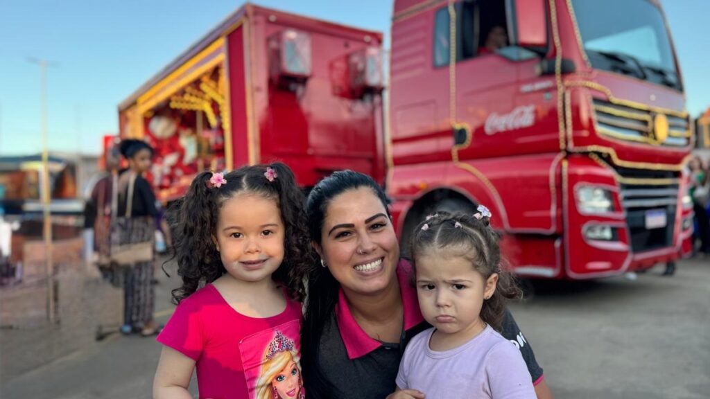 A imagem mostra uma mulher sorridente abraçando duas crianças pequenas em frente a um caminhão vermelho decorado. As crianças parecem ser irmãs, com uma delas sorrindo e a outra com uma expressão um pouco emburrada. O caminhão tem a logo da Coca-Cola e luzes decorativas, sugerindo que pode ser parte de um evento ou promoção. Ao fundo, outras pessoas e estruturas sugerem um ambiente externo, possivelmente uma feira ou evento comunitário. A iluminação sugere que a foto foi tirada durante o dia, com o sol a pino.