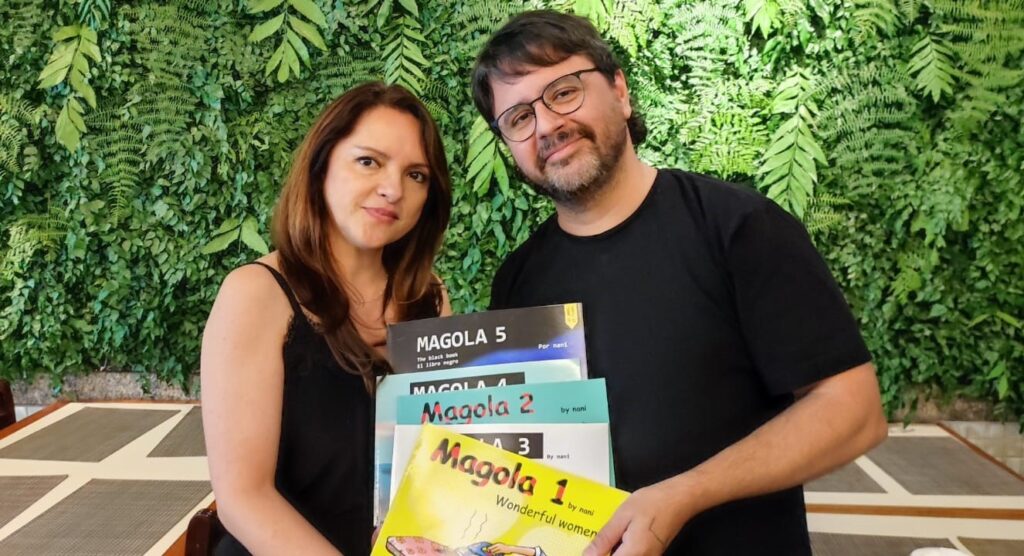 A imagem mostra um homem e uma mulher em pé em frente a uma parede verde coberta de plantas. O homem, à direita, usa óculos e uma camiseta preta. Ele está segurando uma pilha de livros. A mulher, à esquerda, usa uma blusa preta de alcinha e tem cabelos castanhos compridos. Ambos sorriem para a câmera. Os livros que o homem segura são intitulados "MAGOLA 5", "MAGOLA 4", "Magola 2", "Magola 3" e "Magola 1". Alguns dos livros também têm legendas como "The black book", "El libro negro", "by nani" e "Wonderful women". A pilha de livros está posicionada de forma que os títulos sejam visíveis. O fundo da imagem é preenchido com uma parede de folhagens verdes exuberantes, criando um contraste com as pessoas e os livros. Há também uma mesa com um tampo xadrez em tons de bege e cinza visível na parte inferior da imagem.