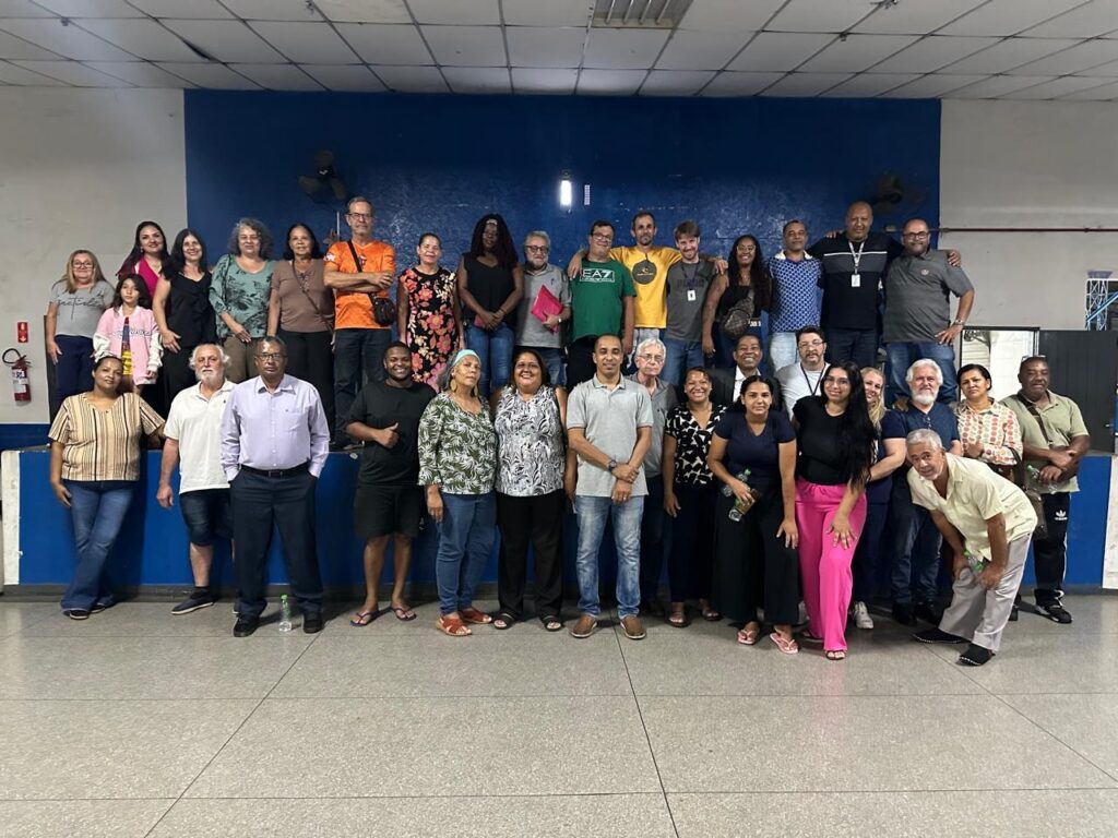 A imagem mostra um grupo de pessoas posando para uma foto, possivelmente após um evento ou reunião. Há 33 pessoas na foto, incluindo homens e mulheres de diversas idades e etnias. Eles estão em pé e sentados em degraus, com um fundo azul escuro e uma parede branca atrás deles. As roupas variam de casuais a mais formais, como camisas sociais e calças. Alguns membros do grupo estão de braços dados, sugerindo um senso de camaradagem. A iluminação é artificial e parece vir de cima, criando algumas sombras no chão. A composição geral da foto é um pouco informal, com algumas pessoas olhando diretamente para a câmera e outras conversando entre si.