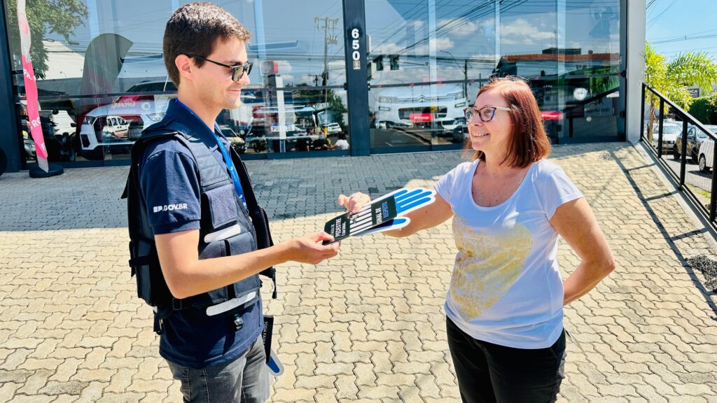 A imagem mostra um homem e uma mulher em frente a uma loja com fachada de vidro. O homem, vestindo uma camiseta azul com o logo "SP.GOV.BR" e um colete tático, está entregando um folheto em formato de mão para a mulher. O folheto tem as cores azul e branca e contém a inscrição "FAIXA DE PEDESTRE" e "SINAL DE RESPEITO". A mulher, usando óculos e uma camiseta branca com uma estampa dourada em formato de coração, sorri enquanto recebe o folheto. Ao fundo, através do vidro da loja, é possível ver carros expostos e o número 650 na fachada. O chão é pavimentado com blocos de concreto intertravados.