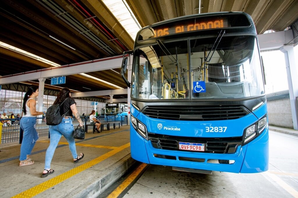 A imagem mostra a frente de um ônibus azul em uma rodoviária. O ônibus tem o número 32837 visível na parte inferior da frente, e o texto "Prefeitura de Piracicaba" também está presente. Acima do para-brisa, um letreiro digital exibe a rota "106 ALGODOAL". Um símbolo azul de acessibilidade para cadeirantes está fixado no vidro do motorista. Ao lado do ônibus, duas mulheres estão caminhando na plataforma. A mulher mais próxima do ônibus usa jeans, uma camiseta preta e carrega uma mochila preta e uma bolsa. A outra mulher, mais distante, usa jeans e uma camiseta branca, e carrega uma bolsa. Ambas usam sandálias. Ao fundo, outros ônibus azuis estão estacionados e pessoas estão sentadas em bancos azuis na área de espera. A rodoviária possui uma cobertura de metal e iluminação artificial. O chão da plataforma é marcado com linhas amarelas de segurança.