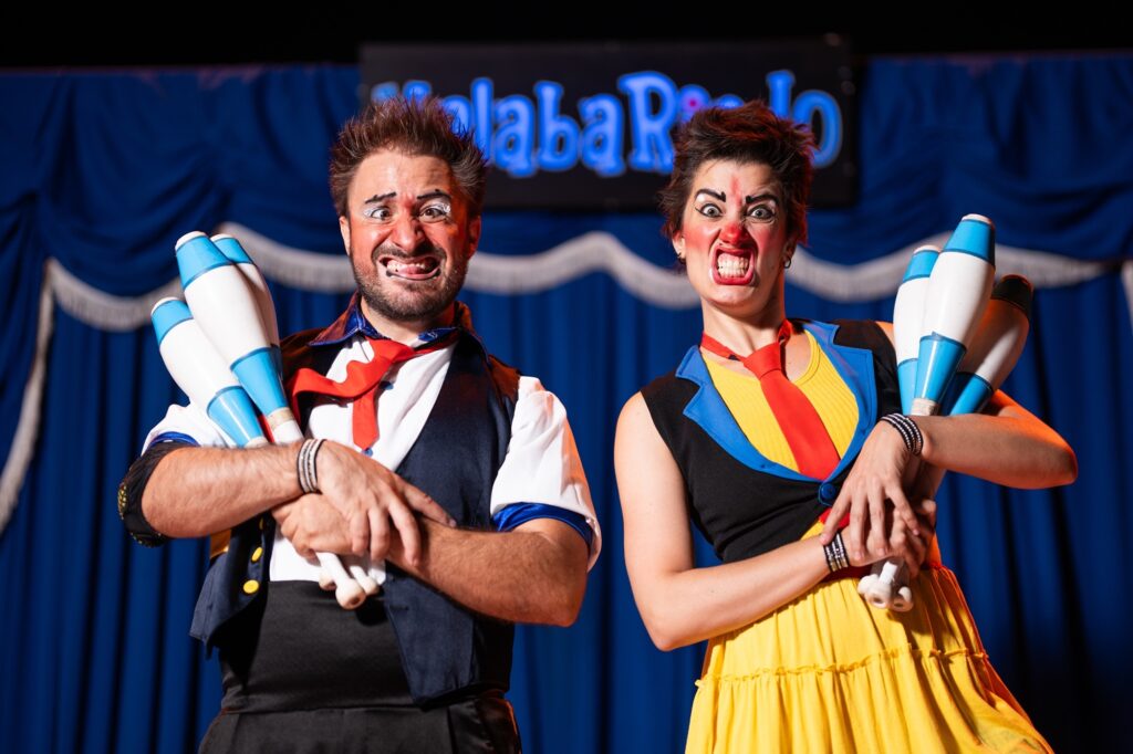 A imagem mostra dois palhaços, um homem e uma mulher, em um palco com um fundo azul drapeado. Ambos estão usando maquiagem de palhaço exagerada, com os rostos expressando uma mistura de alegria e travessura. O palhaço homem, à esquerda, tem cabelo castanho despenteado e um bigode. Ele está usando uma camisa branca com um colete azul escuro e uma gravata vermelha. Ele segura um conjunto de três pinos de malabarismo brancos e azuis em seus braços cruzados. Sua boca está aberta em um sorriso largo, mostrando os dentes, e seus olhos estão arregalados. A palhaça mulher, à direita, tem cabelo escuro curto e usa um colete preto com detalhes azuis e amarelos sobre uma blusa amarela. Ela também usa uma gravata vermelha. Ela segura um conjunto de pinos de malabarismo semelhantes aos do homem. Sua expressão facial é igualmente exagerada, com a boca aberta em um rosnado e os olhos bem abertos, transmitindo uma energia cômica.