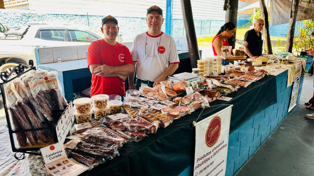 A imagem mostra uma barraca de feira com uma variedade de carnes curadas e defumadas, além de queijos e outros produtos artesanais. Dois homens, um vestindo uma camiseta vermelha e outro uma camiseta branca com o logo "Gianni", estão atrás da barraca, sorrindo para a câmera. A barraca está coberta com uma toalha preta e exibe diversos produtos embalados a vácuo, como linguiças, salames, costelas e outros cortes de carne. Há também potes transparentes com o que parecem ser torresmos ou outros petiscos fritos. Em destaque, há um folheto com a imagem de "Calabresa Curada" e informações sobre os produtos da marca "Gianni". Ao fundo, é possível ver outras pessoas interagindo na feira, incluindo uma mulher e um homem mais velho examinando os produtos em uma outra seção da barraca, onde há queijos e geleias. Uma picape branca está estacionada atrás da barraca, e ao fundo, um muro azul e estruturas de madeira indicam que o evento ocorre ao ar livre ou em um espaço aberto. A atmosfera geral é de um mercado local ou feira de produtores.