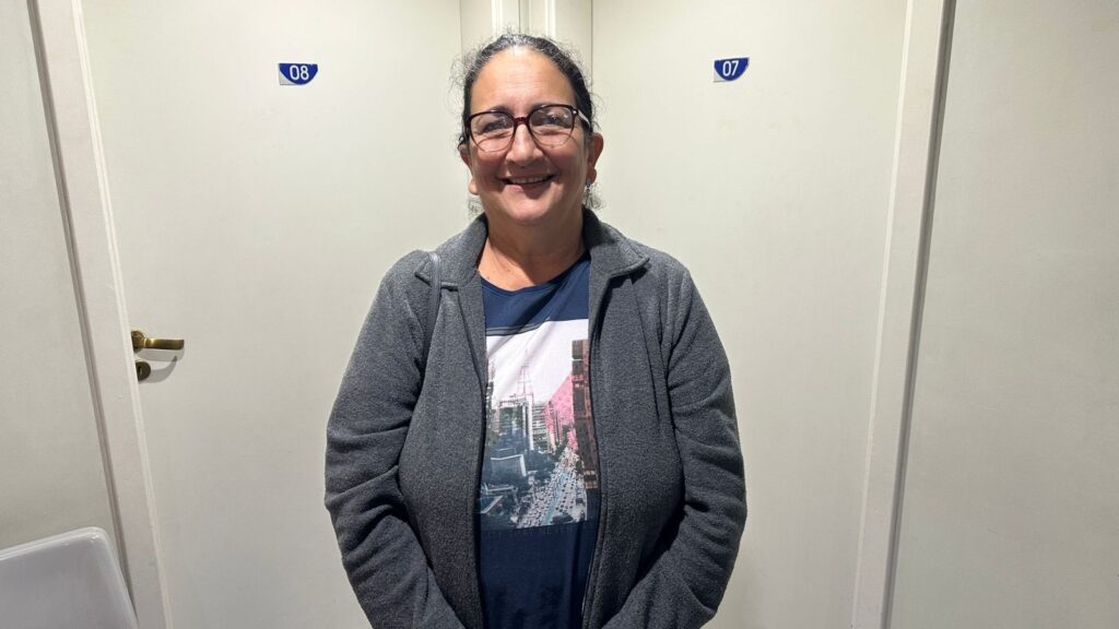 A mulher está sorrindo para a câmera, usando óculos e uma jaqueta cinza sobre uma camiseta azul com uma estampa de uma cidade. Ela está em frente a uma parede branca com duas portas ao lado dela. Uma porta à esquerda tem uma maçaneta dourada e um sinal azul com o número "08" afixado acima dela. Uma porta à direita tem um sinal azul com o número "07" afixado acima dela.