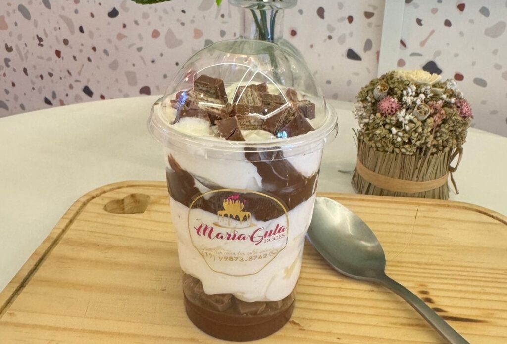 A imagem mostra um copo de sobremesa de chocolate e creme, com pedaços de chocolate e biscoito por cima, em um copo plástico transparente com tampa. O copo está sobre uma tábua de madeira clara com um pequeno entalhe em forma de coração. Ao lado do copo, há uma colher de metal. Ao fundo, há um pequeno arranjo de flores secas e um vaso de vidro com plantas. A parede ao fundo tem um padrão de salpicos em tons de bege, rosa e cinza. O copo tem um rótulo com o nome "Maria Gula Doces" e um número de telefone.