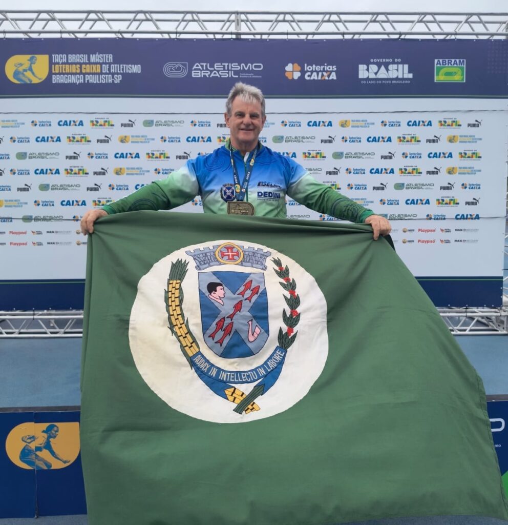 O homem na foto está segurando uma bandeira verde com um brasão ao centro. Ele está sorrindo e vestindo uma camisa de manga comprida com cores azul e verde, além de medalhas no pescoço. Ao fundo, há um banner com a inscrição "TAÇA BRASIL MÁSTER LOTERIAS CAIXA DE ATLETISMO BRAGANÇA PAULISTA-SP" e vários logotipos de patrocinadores como CAIXA e PUMA. A bandeira que ele segura tem um brasão circular com um homem, peixes e uma engrenagem, além de uma inscrição em latim na parte inferior.