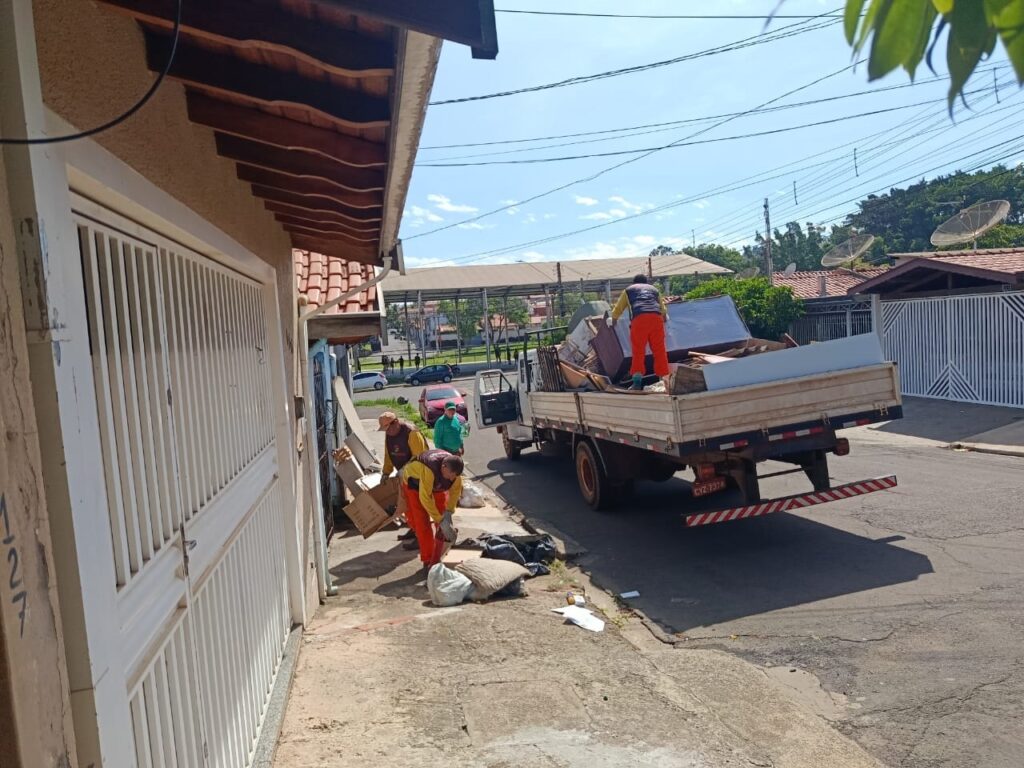 A imagem mostra trabalhadores uniformizados, vestindo coletes laranjas e calças, empenhados na remoção de entulho e móveis de uma residência. Um caminhão de carga está estacionado na rua, com a caçamba repleta de itens descartados. Dois trabalhadores estão próximos à calçada, manipulando caixas e sacos, enquanto um terceiro está sobre o caminhão, organizando a carga. A cena ocorre em um dia ensolarado, em uma rua residencial com casas e árvores ao fundo.