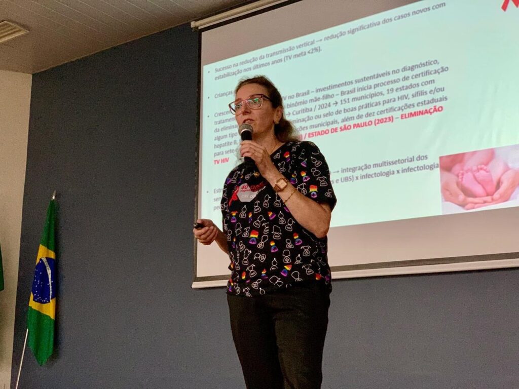 A imagem mostra uma mulher apresentando em um ambiente que parece ser uma conferência ou palestra. Ela está de pé, segurando um microfone com a mão direita e gesticulando com a esquerda. Ela usa óculos e uma blusa preta com estampa colorida, possivelmente um uniforme relacionado à área da saúde. Ao seu lado, há uma tela de projeção exibindo um slide com texto em português, que fala sobre o sucesso na redução da transmissão vertical de doenças como HIV, sífilis e hepatite B, além de mencionar metas de eliminação e certificações em municípios e estados do Brasil. No fundo, há uma parede escura e uma bandeira do Brasil em um suporte, indicando que o evento provavelmente ocorre no Brasil. A iluminação destaca a apresentadora e a tela, enquanto o restante do ambiente está mais escuro. A imagem transmite um contexto de apresentação informativa, possivelmente relacionada à saúde pública e programas de prevenção.