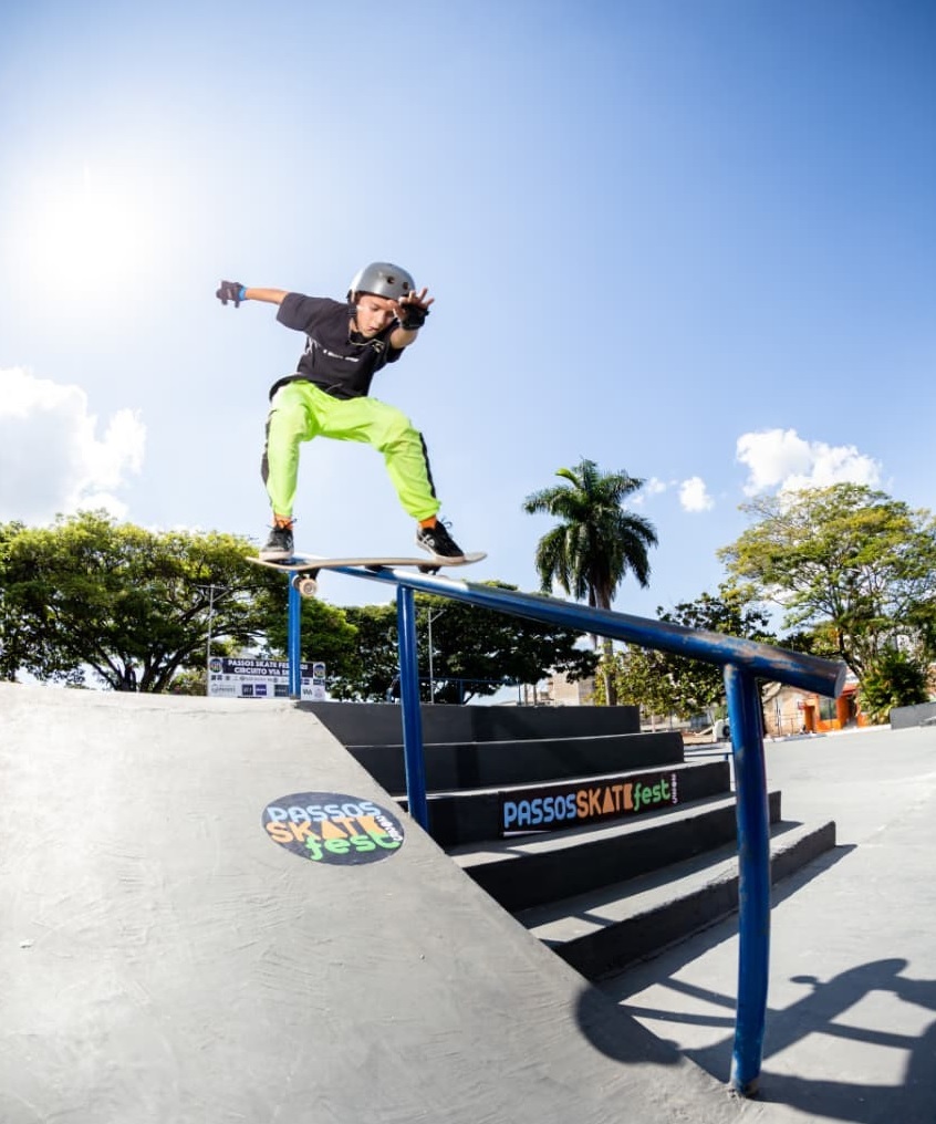 O skatista, usando capacete e calças neon, desliza habilmente sobre um corrimão em uma pista de skate sob um céu azul claro. O sol brilha intensamente, criando uma atmosfera vibrante. Ao fundo, árvores frondosas e uma palmeira adicionam um toque tropical à cena. A rampa de skate tem um adesivo circular com o logo "PASSOS SKATE fest" e uma escadaria com o mesmo nome em destaque.