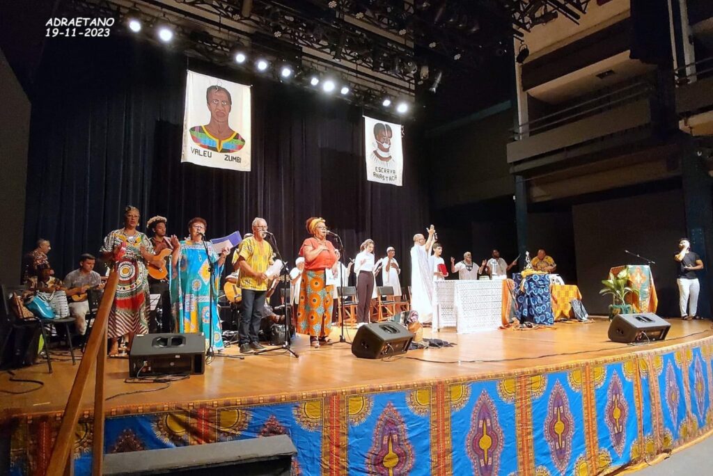 A imagem mostra um grupo de pessoas em um palco, aparentemente durante uma apresentação. Há duas bandeiras penduradas no fundo, com retratos e escritos em português. As pessoas no palco estão vestidas com roupas coloridas e vibrantes, algumas segurando instrumentos musicais como violões e outros objetos, e parecem estar cantando ou falando em microfones. O palco é decorado com um tecido estampado em cores vivas, com um padrão que remete à cultura africana. Há iluminação de palco direcionada para os artistas. Ao fundo, pode-se ver a estrutura de um teatro ou auditório, com assentos e uma varanda. As bandeiras no fundo têm os dizeres "VALEU ZUMBI" e "ESCRAVA ANASTÁCIA", sugerindo uma temática relacionada à história e cultura afro-brasileira. A data "ADRAETANO 19-11-2023" está escrita no canto superior esquerdo da imagem, indicando o local e a data do evento.