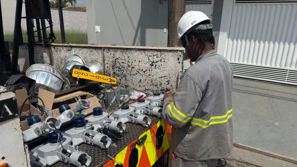 A imagem mostra um trabalhador com um capacete branco e uniforme cinza, em pé ao lado de um caminhão. O caminhão está carregado com vários componentes elétricos, incluindo várias luminárias de poste cinzas com tampas azuis e fios expostos, bem como algumas peças metálicas brilhantes, possivelmente refletores. Um trabalhador está concentrado, examinando ou manipulando um dos componentes. O fundo sugere um ambiente externo, com uma parede cinza e vegetação ao longe.