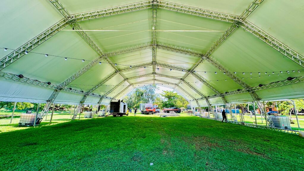 A imagem mostra o interior de uma grande tenda ou pavilhão, com uma estrutura metálica aparente e um teto de lona translúcida, tingindo o ambiente com uma luz verde suave. O chão é coberto por grama verde bem aparada, e ao fundo, árvores e céu azul indicam um ambiente externo. A estrutura do pavilhão é composta por treliças metálicas que formam um arco imponente, sustentando a cobertura. Ao longo das treliças, estão penduradas luzes em formato de lâmpadas, que adicionam um toque decorativo ao espaço. No chão, ao longo das laterais da tenda, observam-se alguns objetos, como recipientes grandes e brancos (possivelmente tanques de água ou material de construção) e caminhões, sugerindo que o local está em fase de montagem ou preparação para um evento. Há também algumas pessoas circulando pelo espaço, indicando atividade e movimento.