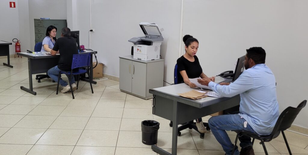Na mesa da frente, uma mulher com blusa preta e cabelo preso em um coque está sentada, entregando papéis para um homem com camisa azul clara e jeans. Ao lado da mesa, há uma lixeira preta. Um pouco mais atrás, em outra mesa, uma mulher com blusa azul clara está sentada em frente a um computador, conversando com um homem com camisa preta e jeans. Atrás deles, há um arquivo de metal cinza. No fundo, entre as duas mesas principais, há uma impressora multifuncional sobre um armário cinza. O chão do escritório é revestido com azulejos quadrados de cor creme. Há também uma extintor de incêndio vermelho na parede, perto da primeira mesa.