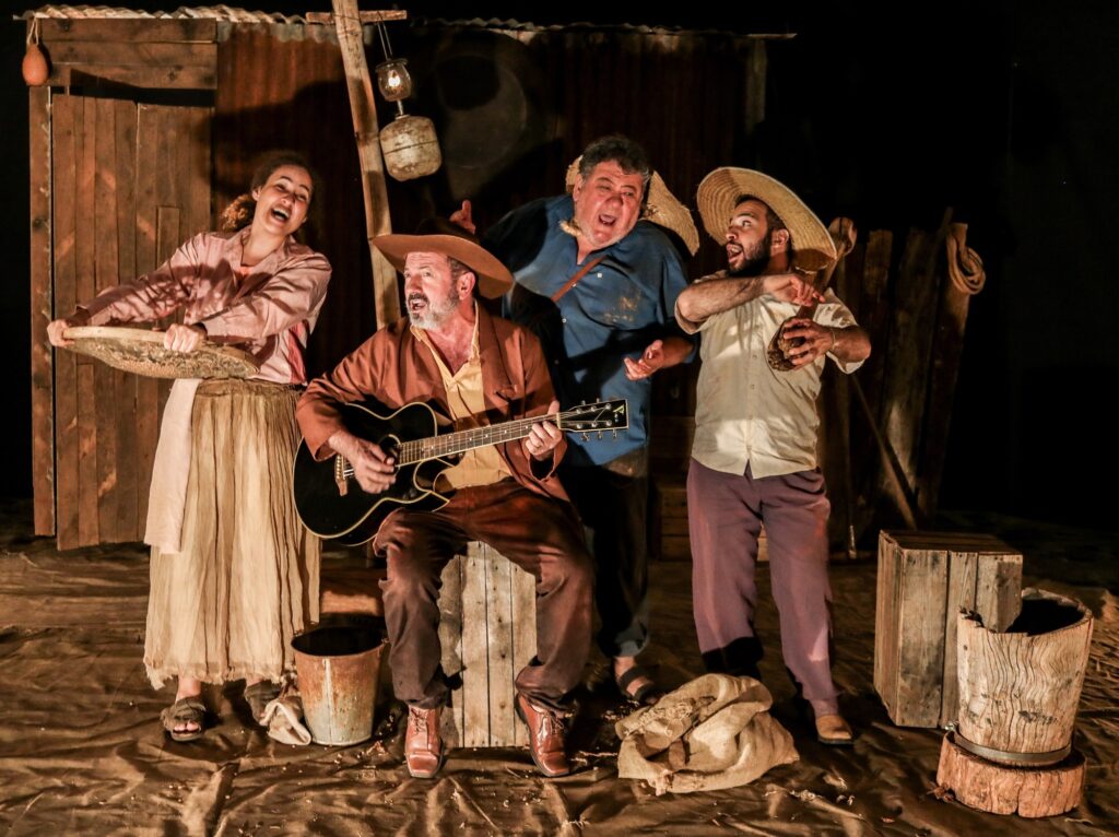 A imagem retrata uma cena teatral com cinco atores em um palco rústico, que simula um ambiente rural ou histórico. Ao centro, um homem toca violão, vestido com uma camisa amarela e um casaco marrom, sentando-se em uma caixa de madeira. À sua esquerda, uma mulher com um vestido longo e claro e uma blusa rosa segura uma espécie de peneira ou cesto, com uma expressão facial animada. À direita do guitarrista, um homem com camisa azul e expressão vocal intensa gesticula com as mãos, usando um chapéu de palha. Ao lado dele, um homem com chapéu de palha semelhante, camisa clara e calças roxas, segura um objeto escuro nas mãos e parece cantar ou gritar. Um quinto homem, parcialmente visível à direita, também usa um chapéu de palha e está em pé. O cenário é composto por tábuas de madeira escuras ao fundo, simulando uma estrutura precária. Objetos como baldes enferrujados, sacos de estopa e toras de madeira completam a ambientação, sugerindo um contexto de trabalho rural ou de vida simples. A iluminação foca nos atores, criando um contraste com o fundo mais escuro.