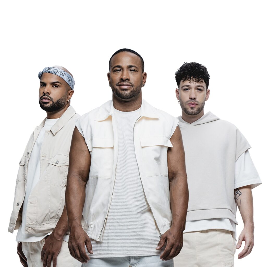 A imagem mostra três homens em pé, em um fundo branco. O homem do lado esquerdo está usando um bandana cinza com estampa paisley na cabeça, uma camisa branca e um colete bege. Ele tem barba e um brinco em uma orelha. O homem do meio está no centro da imagem, vestindo uma camiseta branca texturizada e um colete branco sem mangas com zíper. Ele tem uma barba curta e usa brincos em ambas as orelhas. O homem do lado direito está vestindo uma camiseta branca e um moletom bege folgado, com as mangas cortadas. Ele tem cabelo cacheado escuro, barba curta e um brinco em uma orelha. Ele também tem uma tatuagem visível em seu braço. Todos os homens estão vestidos com roupas em tons neutros e claros, como bege e branco, criando uma estética unificada. A iluminação é clara e frontal, destacando os sujeitos contra o fundo branco.