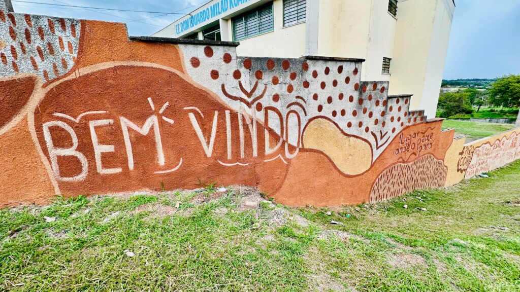 A imagem mostra um muro com arte em relevo e pintura, com a inscrição "BEM VINDO" em letras grandes e brancas sobre um fundo marrom-avermelhado. O muro é decorado com formas abstratas em tons de laranja, amarelo e marrom, e alguns pontos vermelhos sobre um fundo branco na parte superior. À direita, as escadas que sobem o muro também são decoradas com os mesmos padrões. Há uma inscrição em português que diz "Lugar peixe onde o para", com mais texto abaixo, possivelmente nomes de lugares ou frases. Ao fundo, vê-se um edifício escolar com o nome "EE DOM EDUARDO MILAD KOAHAN" escrito na fachada, e um céu nublado. A grama verde e bem aparada cobre o chão em primeiro plano.