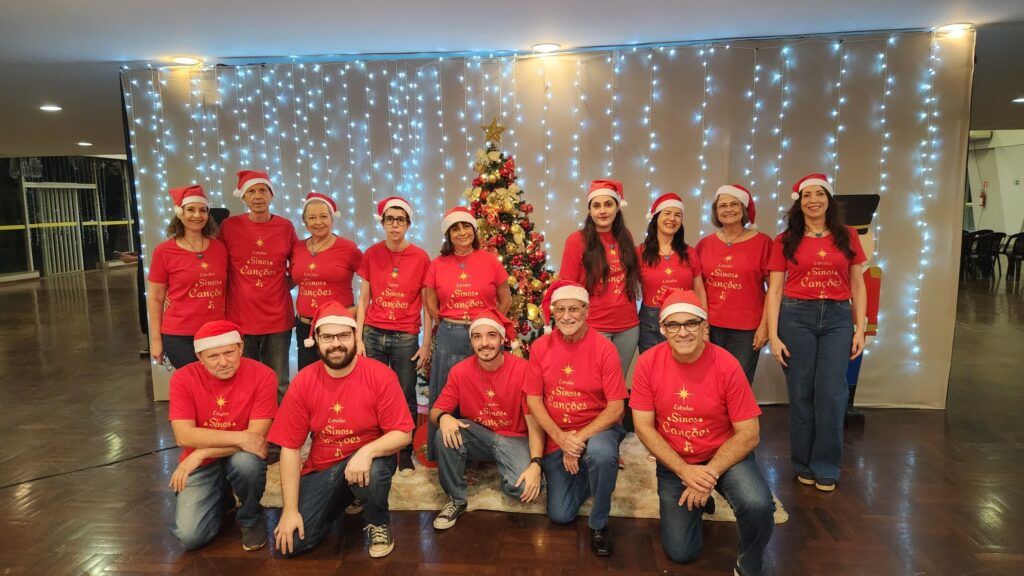 A imagem mostra um grupo de 15 pessoas, 7 homens e 8 mulheres, vestindo camisetas vermelhas com a inscrição "Estrelas Sinos Canções" e chapéus de Papai Noel. Eles estão reunidos em frente a uma árvore de Natal decorada e iluminada por luzes brancas. O grupo está posicionado em duas fileiras, com alguns membros ajoelhados na frente e outros em pé atrás. O fundo é composto por uma cortina branca com fileiras de luzes brancas, criando uma atmosfera festiva de Natal. As pessoas parecem estar celebrando juntas, possivelmente como um coral ou grupo de amigos.