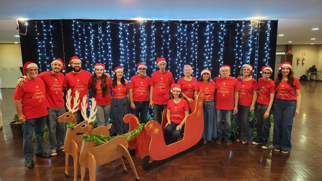 A imagem mostra um grupo de 14 pessoas, 8 homens e 6 mulheres, posando para uma foto em frente a um fundo de luzes de Natal azuis. Todos estão vestindo chapéus de Papai Noel e camisetas vermelhas com a mesma estampa: "Estrelas Sinos Canções" com um desenho de estrela e notas musicais. Na frente do grupo, há um trenó vermelho decorado com estrelas douradas e duas renas de madeira com enfeites verdes de Natal. Uma mulher está sentada dentro do trenó. O grupo parece estar celebrando o Natal ou um evento temático de fim de ano. O piso é de madeira escura e polida. Ao fundo, à direita, há uma porta e um homem sentado em uma cadeira.