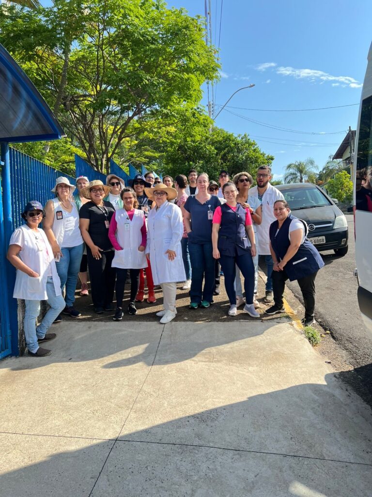 A imagem mostra um grupo de pessoas, a maioria mulheres, posando para uma foto em frente a uma cerca azul e árvores verdes. Algumas pessoas usam jalecos brancos, sugerindo que podem ser profissionais de saúde ou estudantes da área. Outras usam uniformes azuis e escuros, possivelmente de uma equipe de apoio ou outra função profissional. O grupo parece estar em um dia ensolarado, com o céu azul visível acima. Há um carro escuro e parte de um veículo branco (possivelmente uma van ou ônibus) visíveis na lateral da imagem, indicando que eles podem ter chegado ou estão prestes a partir. A postura e as expressões faciais da maioria das pessoas sugerem um momento de descontração e união, talvez antes ou depois de uma atividade ou evento.