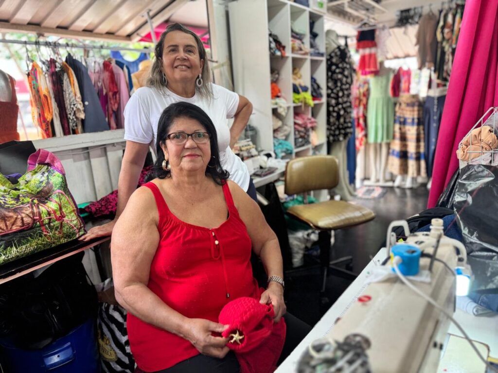 Duas mulheres estão em uma loja de roupas. Uma mulher está sentada em primeiro plano, usando um top vermelho e óculos. Ela está segurando um pedaço de tecido vermelho. Atrás dela, outra mulher está em pé, usando uma camiseta branca. Elas estão em uma loja bem abastecida, com roupas penduradas em araras e prateleiras cheias de itens. Há uma máquina de costura em primeiro plano, sugerindo que a loja pode ser uma alfaiataria ou ter um serviço de costura.