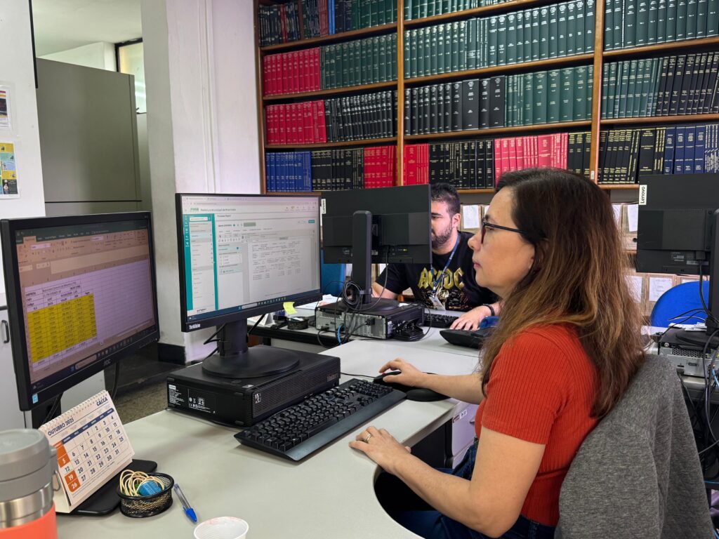 A imagem mostra dois profissionais trabalhando em um escritório. Uma mulher com óculos e uma blusa laranja está sentada em primeiro plano, concentrada em seu computador. Ao fundo, um homem com uma camiseta preta está trabalhando em outro computador. Ambos estão em frente a monitores que exibem informações em tela, possivelmente planilhas ou sistemas. Atrás deles, há uma estante cheia de livros encadernados em cores como vermelho, verde e azul, sugerindo um ambiente de arquivo ou biblioteca. Na mesa da mulher, há um calendário de outubro de 2025, um copo, uma caneta e outros itens de escritório. A iluminação e a organização do espaço indicam um ambiente de trabalho funcional.