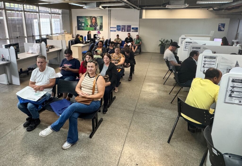 A imagem mostra um interior de escritório com várias pessoas sentadas em cadeiras, aparentemente esperando para serem atendidas. Há mesas com computadores e divisórias, sugerindo um ambiente de serviço ao cliente ou administrativo. Uma grande imagem de uma mulher sorrindo está afixada na parede com o texto "SEBRAE Aqui". As pessoas em espera estão vestidas casualmente, e algumas estão olhando para seus telefones. O ambiente parece limpo e organizado.