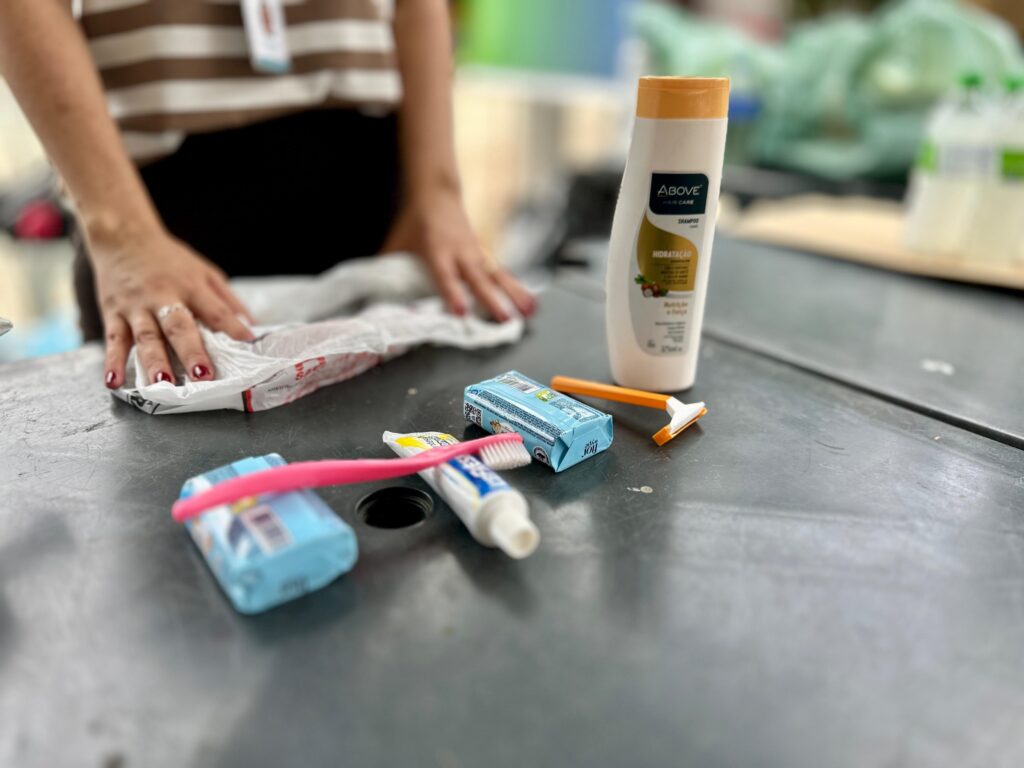 A imagem mostra uma mesa com vários itens de higiene pessoal dispostos sobre ela. Uma pessoa, com as mãos visíveis, está arrumando alguns desses itens. Os itens de higiene pessoal incluem: - Uma garrafa de shampoo da marca "Above Hair Care", com a indicação "Hidratação" e "Nutrição e Força". - Um pequeno sabonete embalado em azul claro. - Uma escova de dentes rosa. - Um tubo de pasta de dentes branco com detalhes azuis e a ponta do tubo visível. - Um aparelho de barbear descartável com cabo laranja e lâmina branca. O fundo da imagem está desfocado, mas é possível identificar um ambiente interno, com alguns objetos em tons de verde e outros itens em garrafas ao fundo. A iluminação sugere um ambiente iluminado, possivelmente durante o dia. A pessoa veste uma blusa listrada em tons de marrom e branco e uma peça preta na parte inferior. Suas unhas estão pintadas de vermelho.