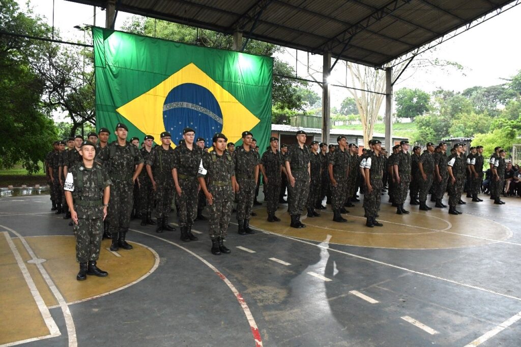A imagem mostra um grupo de jovens em uniforme militar, em pé em formação, em uma quadra coberta. Atrás deles, uma grande bandeira do Brasil está pendurada. Os jovens parecem estar em um evento oficial ou formatura, com a bandeira nacional como pano de fundo solene. O ambiente é ao ar livre, mas coberto, sugerindo um clima tropical. A iluminação é natural, com raios de sol penetrando pela cobertura.