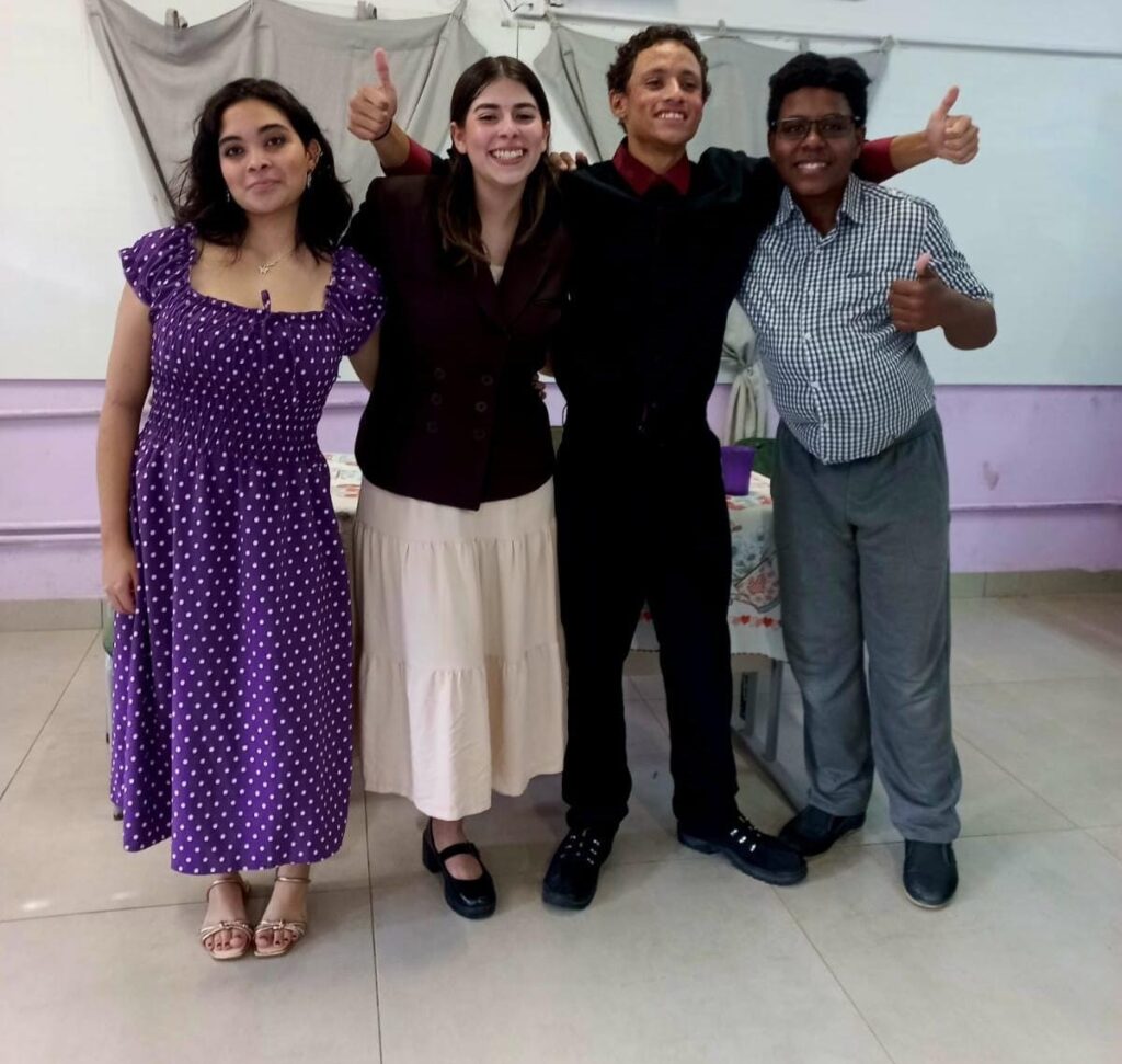 A imagem mostra quatro jovens, duas mulheres e dois homens, posando juntos em um ambiente interno. Eles estão próximos, com os braços ao redor uns dos outros, transmitindo uma sensação de amizade e alegria. Todos sorriem para a câmera, e dois deles fazem sinal de positivo com o polegar. A mulher à esquerda veste um vestido roxo com bolinhas brancas, de mangas curtas e bufantes. Ao seu lado, uma mulher com cabelo castanho claro usa um blazer marrom escuro sobre uma saia clara e rodadinha. O homem ao lado dela usa uma camisa vermelha escura e calça preta, enquanto o homem à direita veste uma camisa xadrez azul e branca e calça cinza. O ambiente parece ser uma sala interna com parede clara e cortina ao fundo. Há uma mesa com toalha estampada e um copo roxo sobre ela, parcialmente visível entre as pessoas. A iluminação é artificial, criando sombras suaves. No geral, a imagem transmite um momento descontraído e feliz entre amigos ou colegas, possivelmente em uma ocasião especial ou evento social.