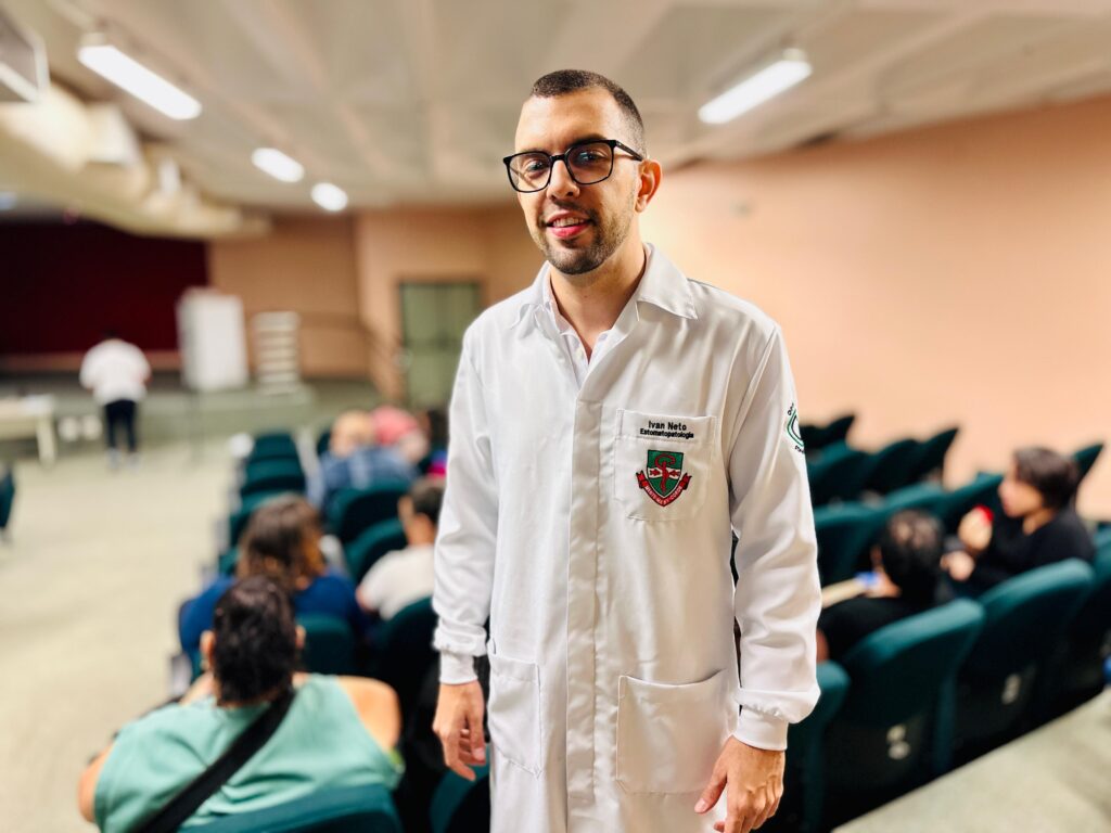 O homem na foto está usando um jaleco branco, que é comumente usado por profissionais de saúde. Ele tem óculos pretos e está sorrindo para a câmera. No jaleco, há um emblema com o nome "Ivan Neto" e a palavra "Estomatopatologia" escrita abaixo. O fundo da imagem mostra uma sala com cadeiras em semicírculo e algumas pessoas sentadas, sugerindo que ele está em uma sala de aula, auditório ou sala de conferências. Há também um palco ou área de apresentação no fundo.
