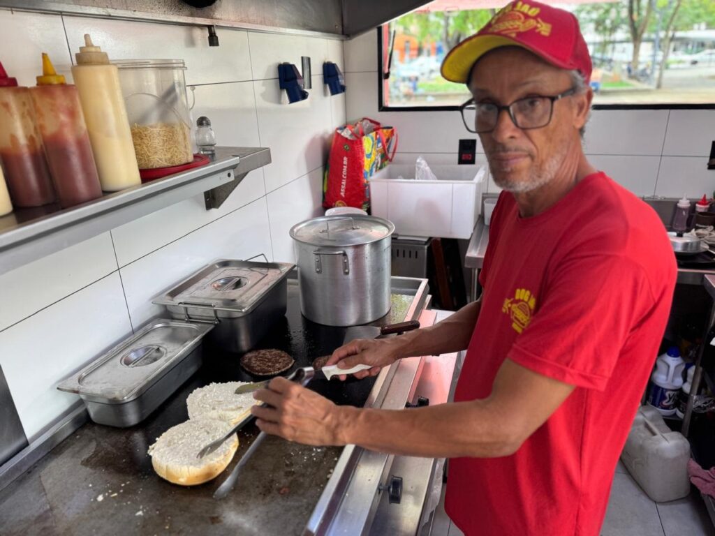 A imagem mostra um homem em uma cozinha, possivelmente um restaurante ou food truck, preparando comida. Ele está usando um boné vermelho e uma camiseta vermelha com um logo. Ele usa óculos e tem um olhar sério enquanto trabalha em uma chapa quente. Na chapa, podemos ver pães de hambúrguer cortados ao meio e alguns hambúrgueres grelhando. Há também recipientes de metal com tampas, que provavelmente contêm ingredientes. Ao fundo, em uma prateleira, há vários frascos de condimentos, incluindo ketchup e maionese, além de um recipiente com batatas fritas finas e um saleiro. A cozinha parece ser um ambiente de trabalho funcional, com paredes revestidas de azulejos brancos.