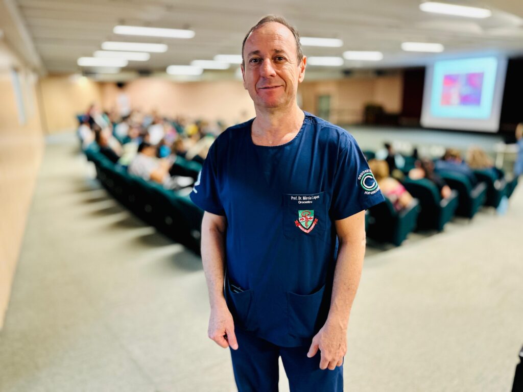 A imagem mostra um homem de meia-idade, vestindo um uniforme azul escuro, possivelmente um scrub médico, em pé em um auditório. Ele está sorrindo para a câmera e tem um crachá preso ao seu uniforme. Atrás dele, um público está sentado em fileiras de assentos verdes, voltados para uma tela de projeção que exibe uma imagem colorida. O ambiente sugere uma palestra, conferência ou apresentação educacional.