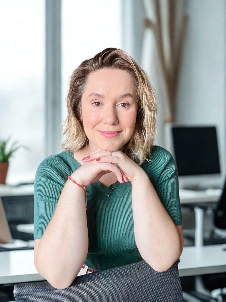A mulher, com cabelo loiro ondulado e pele clara, está sentada em uma cadeira de escritório, com os braços apoiados na cadeira e as mãos unidas sob o queixo. Ela usa uma blusa verde de gola alta com mangas curtas. Um colar fino com um pingente dourado em forma de bola repousa sobre sua blusa. Em seu pulso esquerdo, ela usa uma pulseira vermelha com detalhes dourados. Suas unhas estão pintadas de um tom de marrom avermelhado.O fundo da imagem sugere um escritório moderno, com grandes janelas que deixam entrar luz natural. Vemos mesas de escritório, computadores e uma planta em vaso em um canto. A iluminação suave realça o rosto da mulher, que sorri para a câmera.