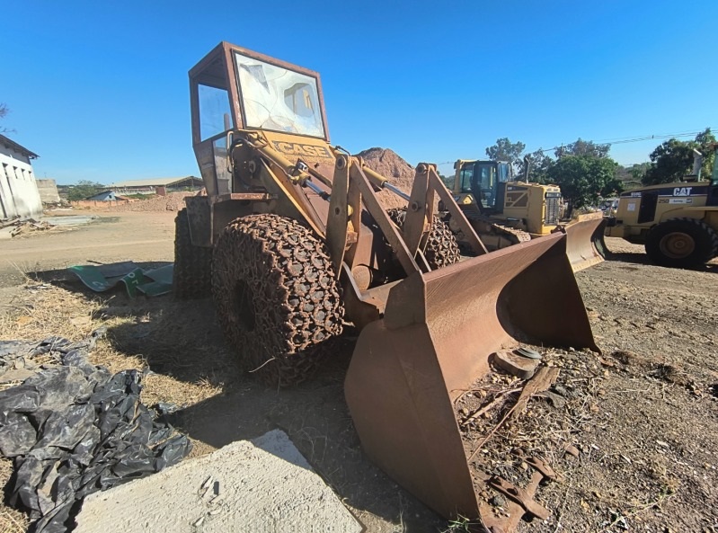 A imagem mostra uma pá carregadeira antiga e enferrujada, com a marca "CASE" visível na lateral. A máquina está em um local a céu aberto, com um céu azul claro ao fundo. Há pilhas de terra ou areia atrás da pá carregadeira e outras máquinas pesadas, como tratores de esteira da marca CAT, ao fundo. A cabine da pá carregadeira tem o vidro quebrado. Há também alguns materiais pretos e cinzas no chão em primeiro plano. A cena sugere um pátio de máquinas ou um local de descarte de equipamentos.