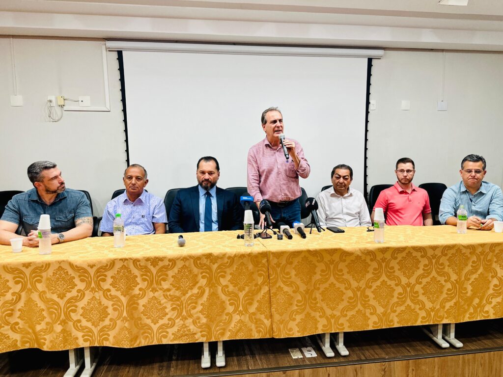 A imagem mostra um grupo de sete homens sentados e um homem em pé, todos atrás de uma mesa longa coberta com uma toalha amarela com um padrão intrincado. O homem em pé, vestindo uma camisa xadrez rosa e jeans, está falando em um microfone. Há vários microfones e garrafas de água sobre a mesa. Os outros seis homens estão sentados em cadeiras pretas, com os braços apoiados na mesa. Cada um deles está vestido de forma formal ou semi-formal, com camisas de botões. O homem mais à esquerda usa uma camisa jeans, enquanto os outros usam camisas de botões em tons de azul, branco e rosa. O homem mais à direita usa uma camisa azul listrada. A sala parece ser uma sala de conferências, com uma tela branca ao fundo. A iluminação é artificial e parece vir de cima.