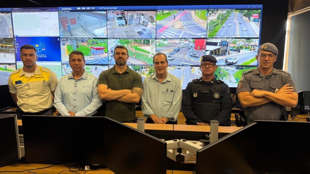 A imagem mostra um grupo de seis homens em pé em frente a uma grande tela de monitoramento composta por vários monitores menores que exibem imagens de câmeras de segurança. Os homens parecem ser funcionários de segurança ou trânsito, pois alguns vestem uniformes. O homem mais à esquerda veste um uniforme amarelo e preto com a palavra "TRANSITO" escrita em seu colete. Ele usa óculos e tem uma barba curta. Os próximos dois homens vestem camisas casuais. O homem do meio tem os braços cruzados e usa uma camiseta verde escura. O homem ao lado dele usa uma camisa xadrez azul e branca com uma caneta no bolso. Os três homens mais à direita vestem uniformes escuros e parecem ser policiais. Eles usam boinas e têm crachás em seus uniformes. Os dois policiais mais à direita têm os braços cruzados. As telas de monitoramento atrás deles exibem uma variedade de imagens de tráfego e ruas, com algumas informações textuais sobrepostas, como "INTELIGENCIA PIRACICABA" e horários. As telas sugerem que este é um centro de controle de tráfego ou segurança.